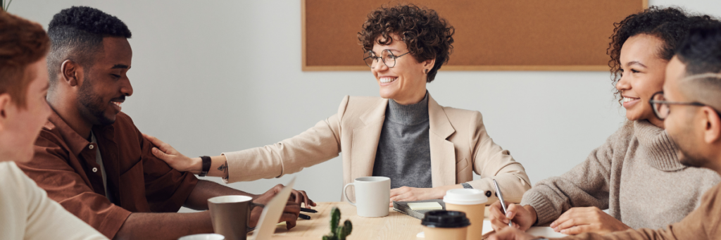 A group of five people around a conference room table having coffee and smiling about something during the meeting