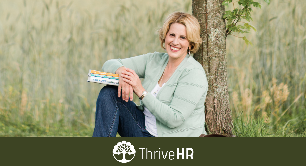 Woman sitting against a tree smiling at the camera holding books encouraging HR Action items