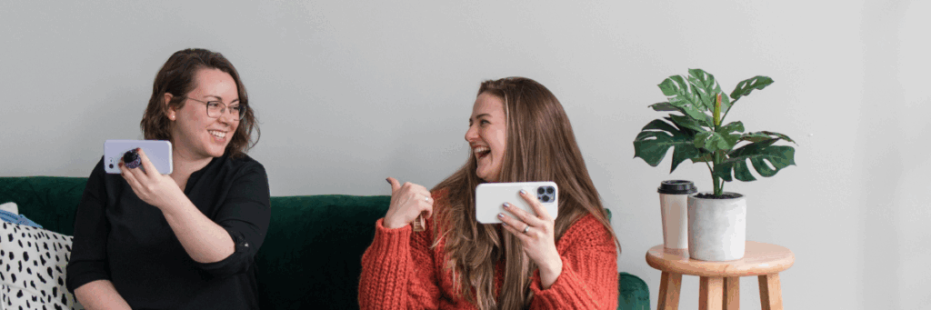 Two women sitting on a couch looking at each other and smiling while holding their phones up In front of them