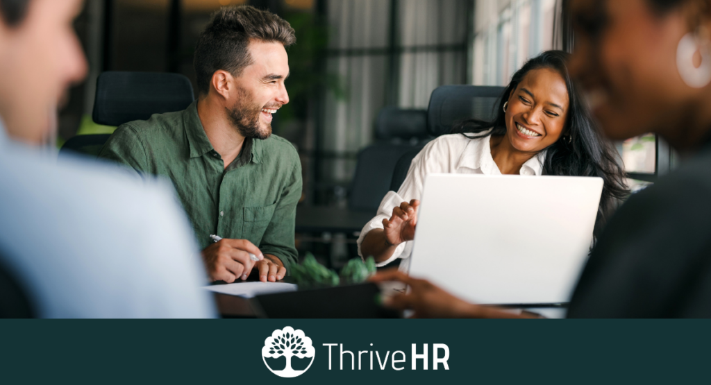 Team members collaborating and laughing around a laptop during an in-person meeting, representing a positive and people-focused workplace culture.